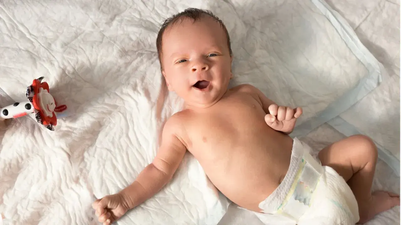 Baby mit offenem Mund, vielleicht im 6-Wochen-Schub, liegt auf einer weißen Unterlage neben einem bunten Spielzeug.