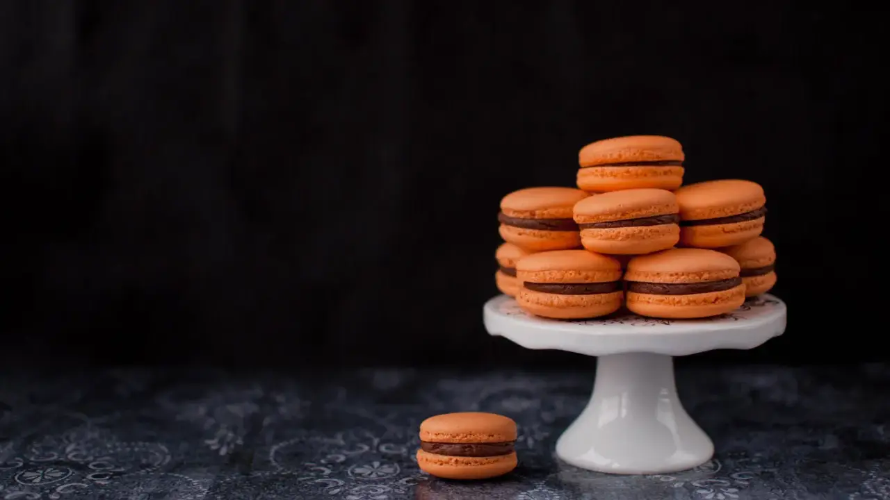 Pomarańczowe makaroniki francuskie z czekoladowym nadzieniem ułożone na białej paterze, jeden makaronik leży obok.