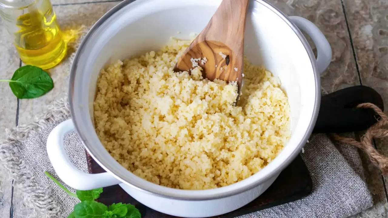 Ein Topf mit lockerem Couscous, bereit für ein marokkanisches Rezept. Ein Holzlöffel rührt die Körner auf. Daneben eine Flasche Öl und frische Minze.