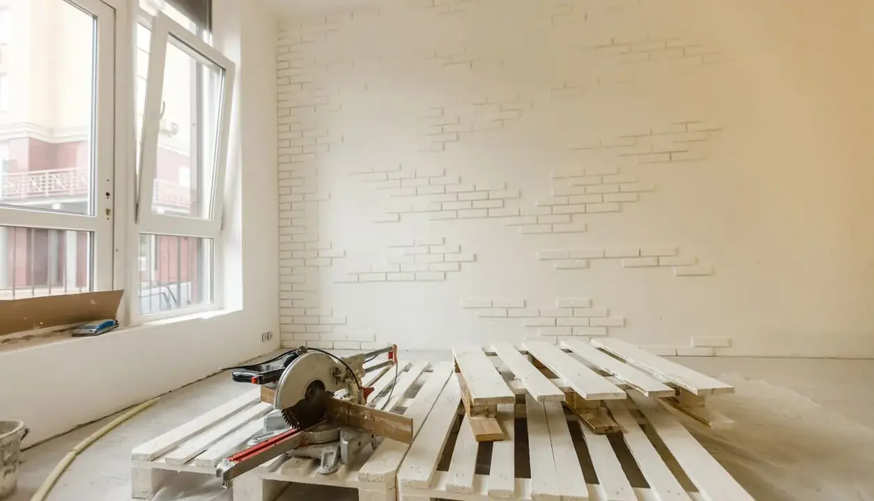 Herramientas listas para como pintar un palet. Madera blanca apilada, sierra ingletadora y pared con ladrillos blancos.