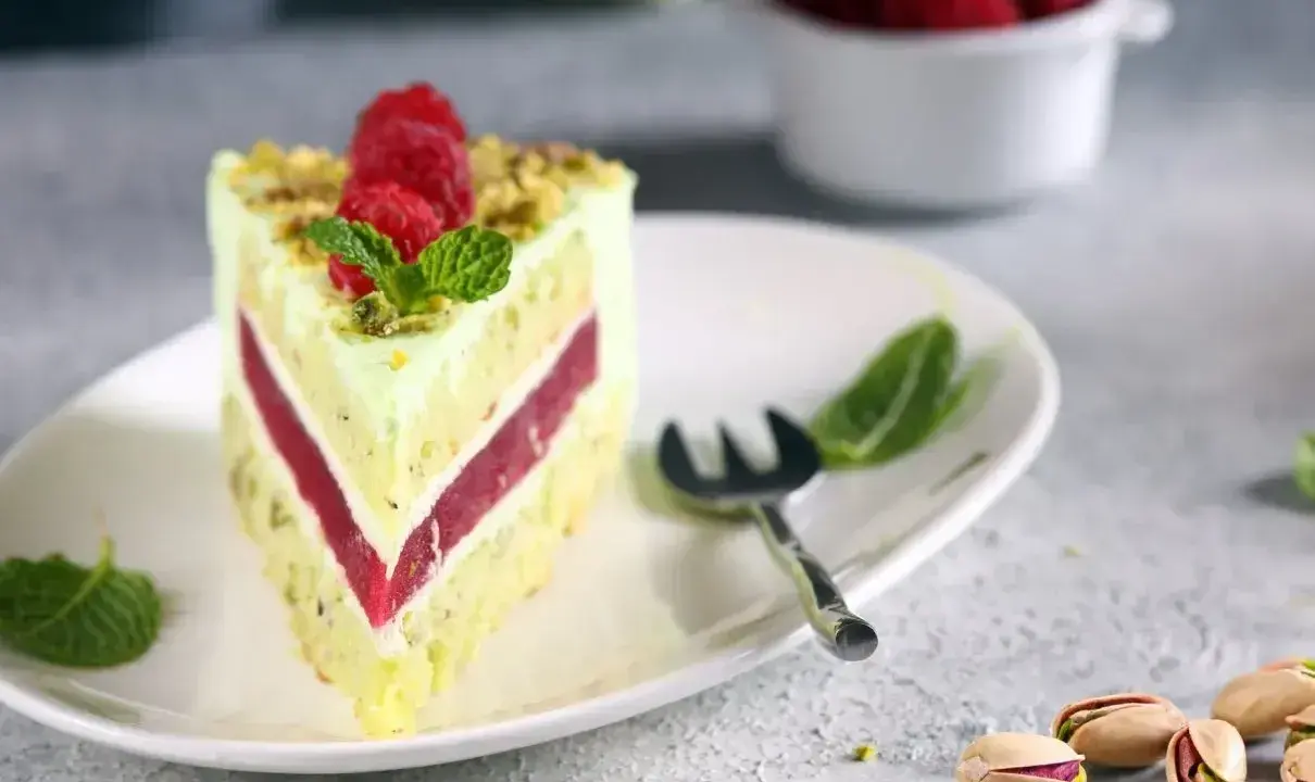 Pyszny tort pistacjowo malinowy z malinami i miętą na talerzu. Obok widoczne łuskane pistacje.