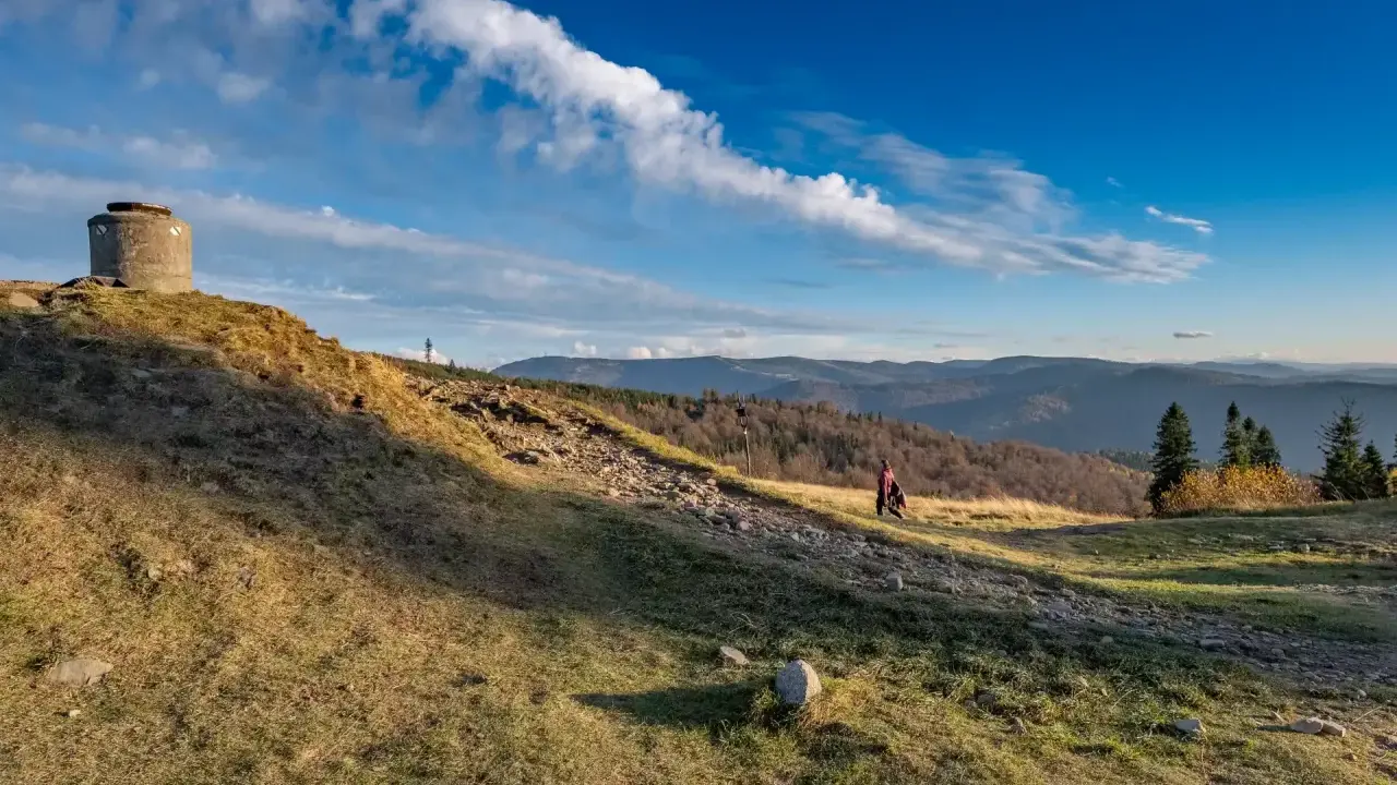 Na zboczu **blatniej góry** stoi betonowy bunkier. W oddali widać pasmo górskie i lasy.