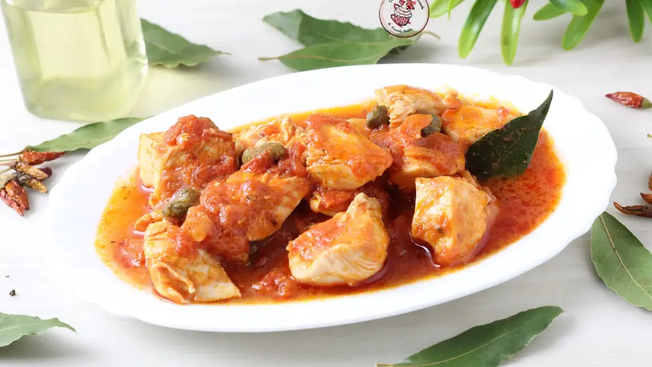 Succulento pollo alla pizzaiola con pomodoro, capperi e olive, guarnito con foglie di alloro e peperoncini secchi.