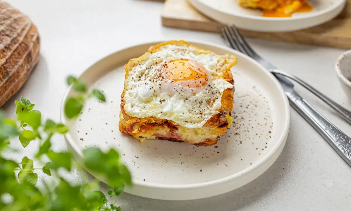 Pyszny croque madame z jajkiem sadzonym na wierzchu, na talerzu, obok chleba i zielonych liści.