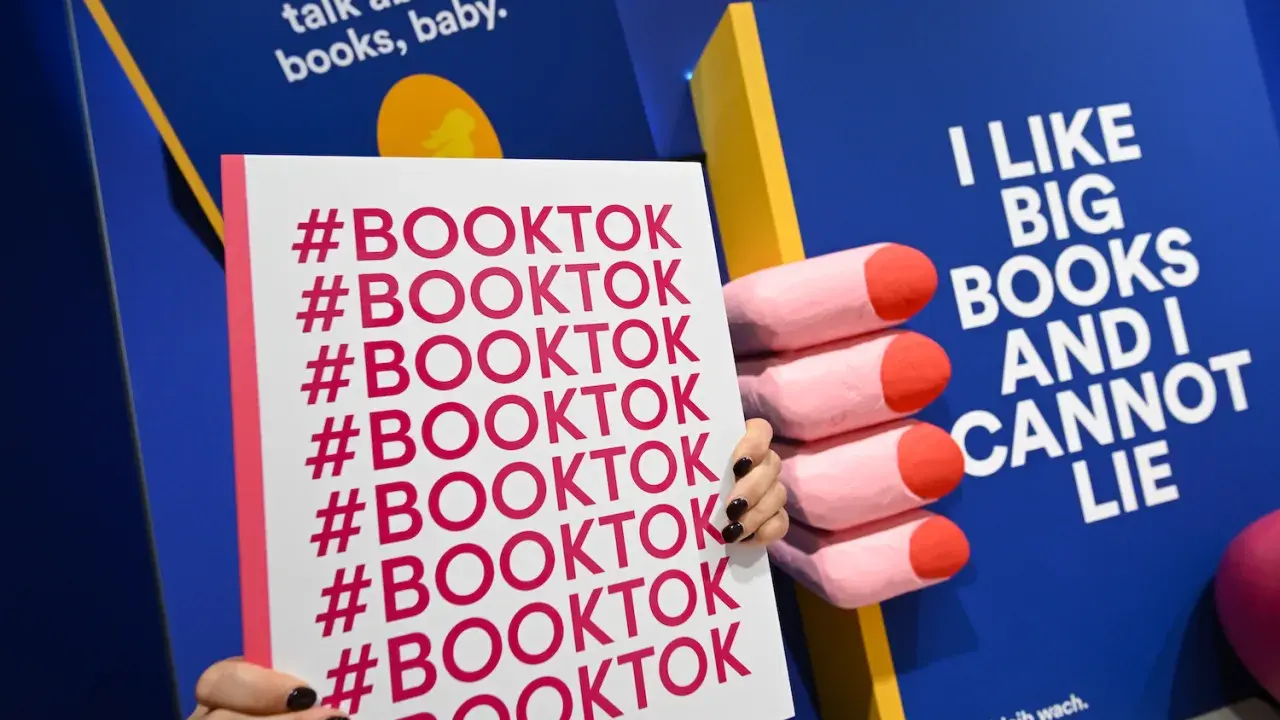 Mani che tengono un cartello con #BOOKTOK ripetuto. Sullo sfondo, un libro blu con la scritta "I LIKE BIG BOOKS AND I CANNOT LIE".