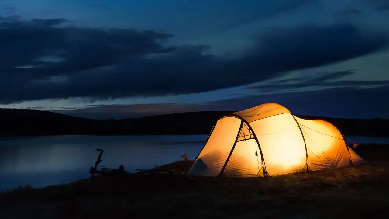 Upplyst tält vid en sjö i skymningen. En cykel står parkerad bredvid. Perfekt för camping Sverige karta-äventyr.