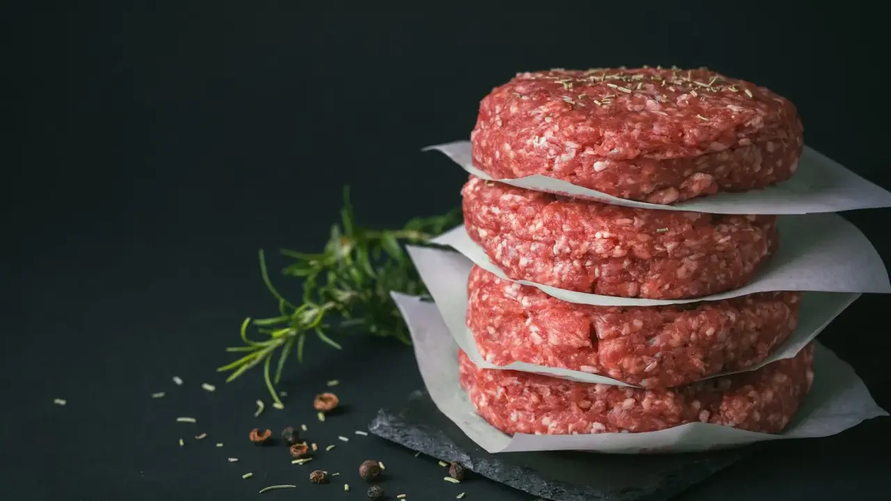Stos świeżego mięsa na burgery, przyprawionego ziołami, obok gałązki rozmarynu i pieprzu.