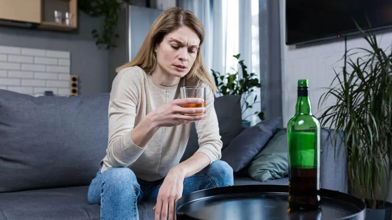 Kobieta z kieliszkiem alkoholu i butelką obok. Obraz symbolizuje 10 strat z picia alkoholu, pokazując jej smutek i zagubienie.