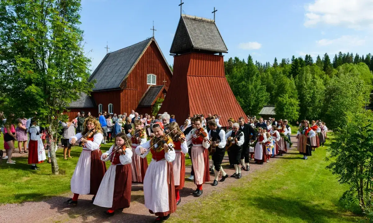 En procession av musiker i folkdräkter, som spelar fiol och gitarr, går förbi en röd träkyrka. Många människor i traditionella kläder, vilka firar midsommar, följer efter.