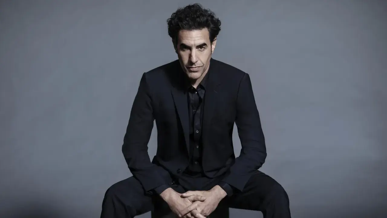 Sacha Baron Cohen, känd för sina kontroversiella filmer, sitter i en mörk kostym med händerna knäppta.