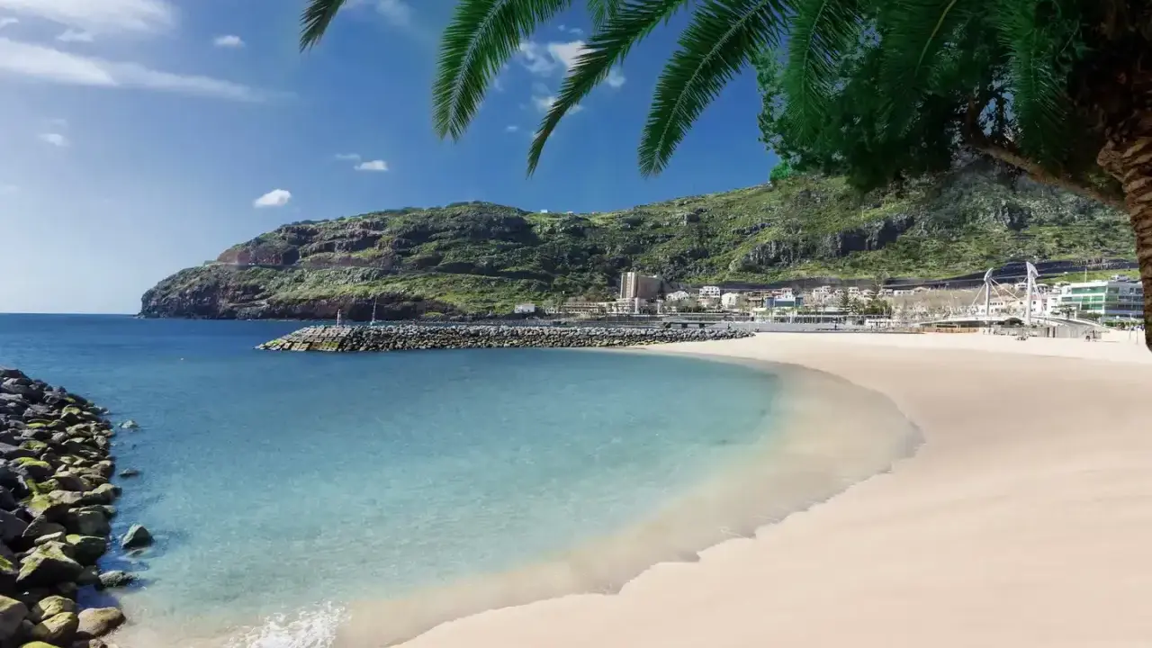Traumhafter Madeira Strand mit türkisfarbenem Wasser, goldenem Sand und grünen Hügeln im Hintergrund. Palmen spenden Schatten.