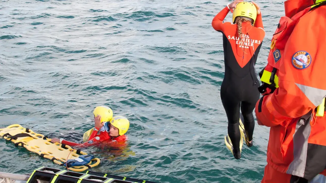 Sauvetage en mer : une sauveteuse en combinaison noire et orange se prépare à plonger. Deux personnes en gilets de sauvetage et casques jaunes flottent près d'une civière. Communication VHF et MMSI essentielle.