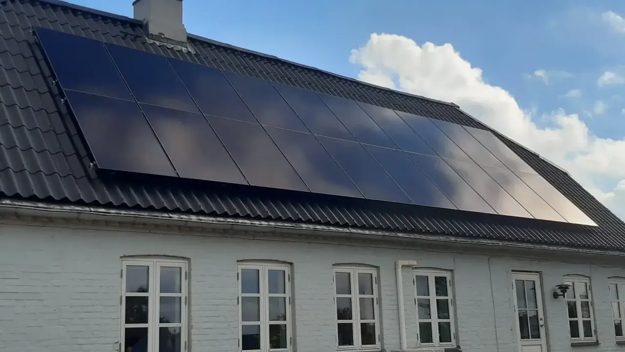 Ett hus med svarta, glänsande nya solceller på taket. Solen reflekteras i panelerna mot en blå himmel med moln.