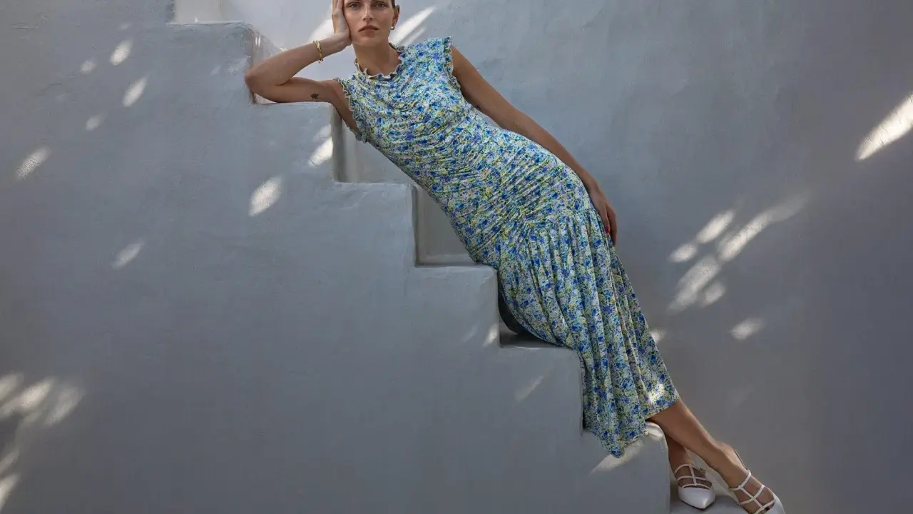 A woman in a floral dress and white heels, embodying a chic formal dress code for women, poses on white stairs.