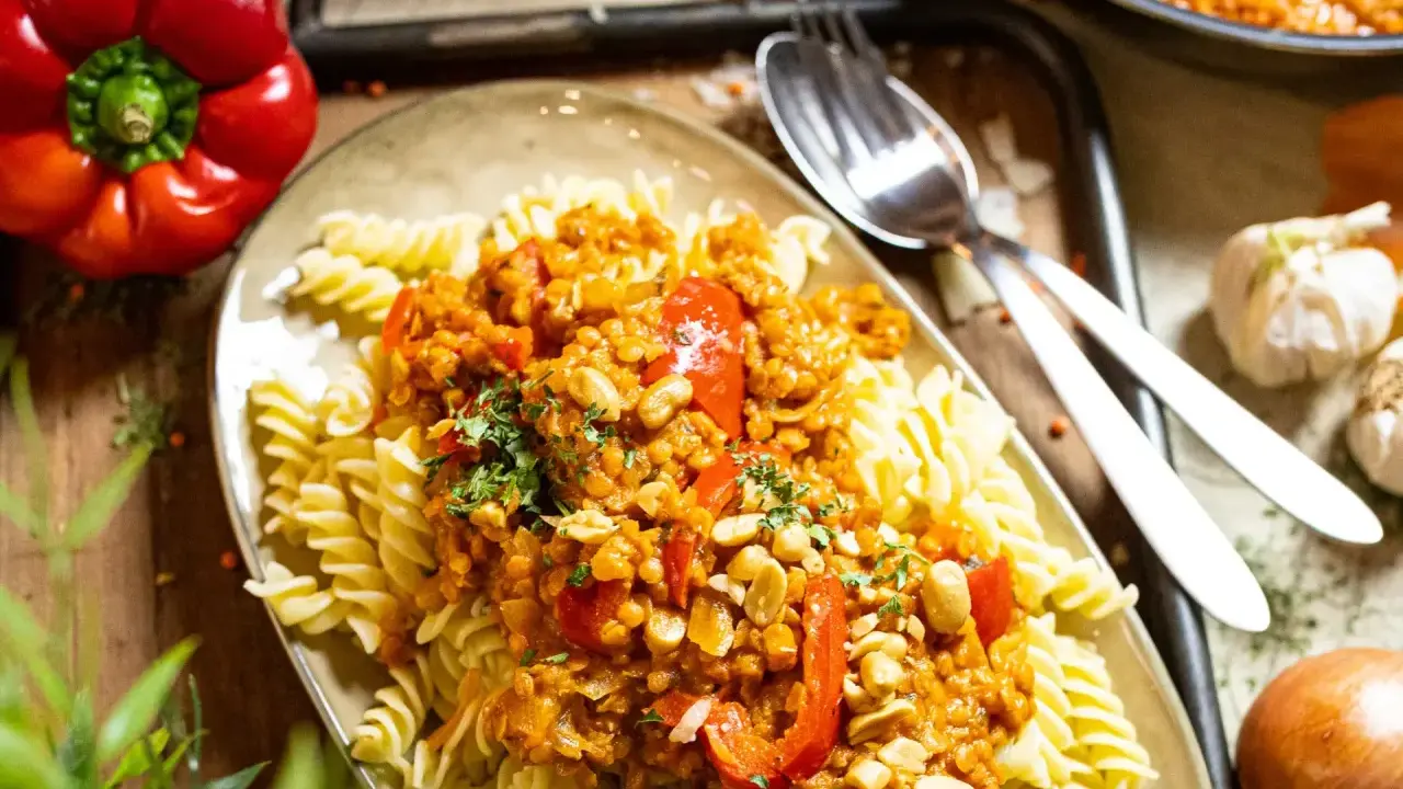 Fusilli mit einer herzhaften bolognese mit roten linsen, garniert mit Erdnüssen und Petersilie. Daneben liegen eine rote Paprika und Knoblauch.
