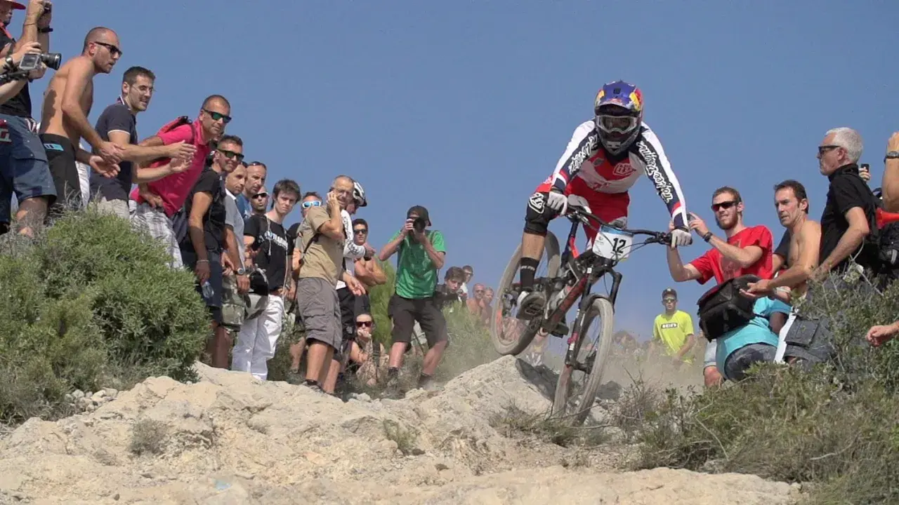 Kolarz w akcji podczas zawodów MTB, otoczony przez kibiców i fotografów.