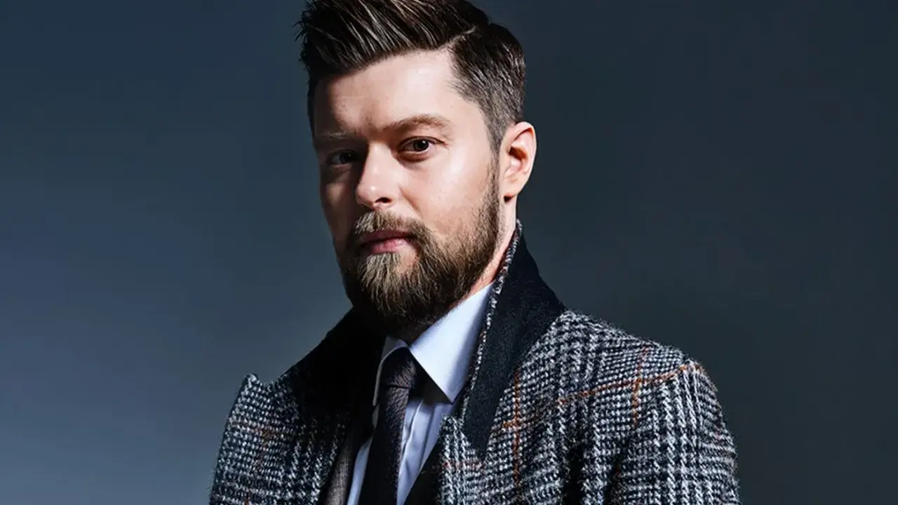 Mężczyzna z brodą i starannie ułożonymi włosami, w eleganckim płaszczu w kratę. Remigiusz Mróz wiek nie jest widoczny, ale styl sugeruje dojrzałość.