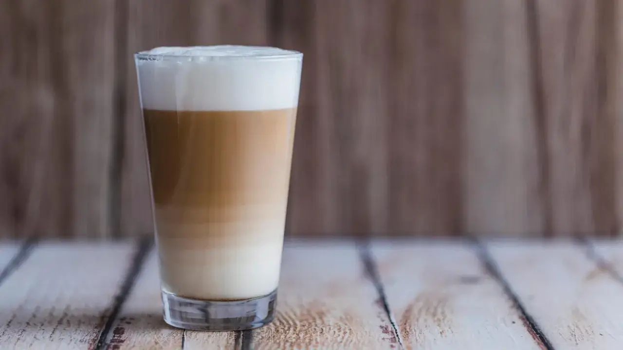 Ein hoher Latte Macchiato mit Mandelmilch, geschichtet in drei Farben, steht auf einem Holztisch.