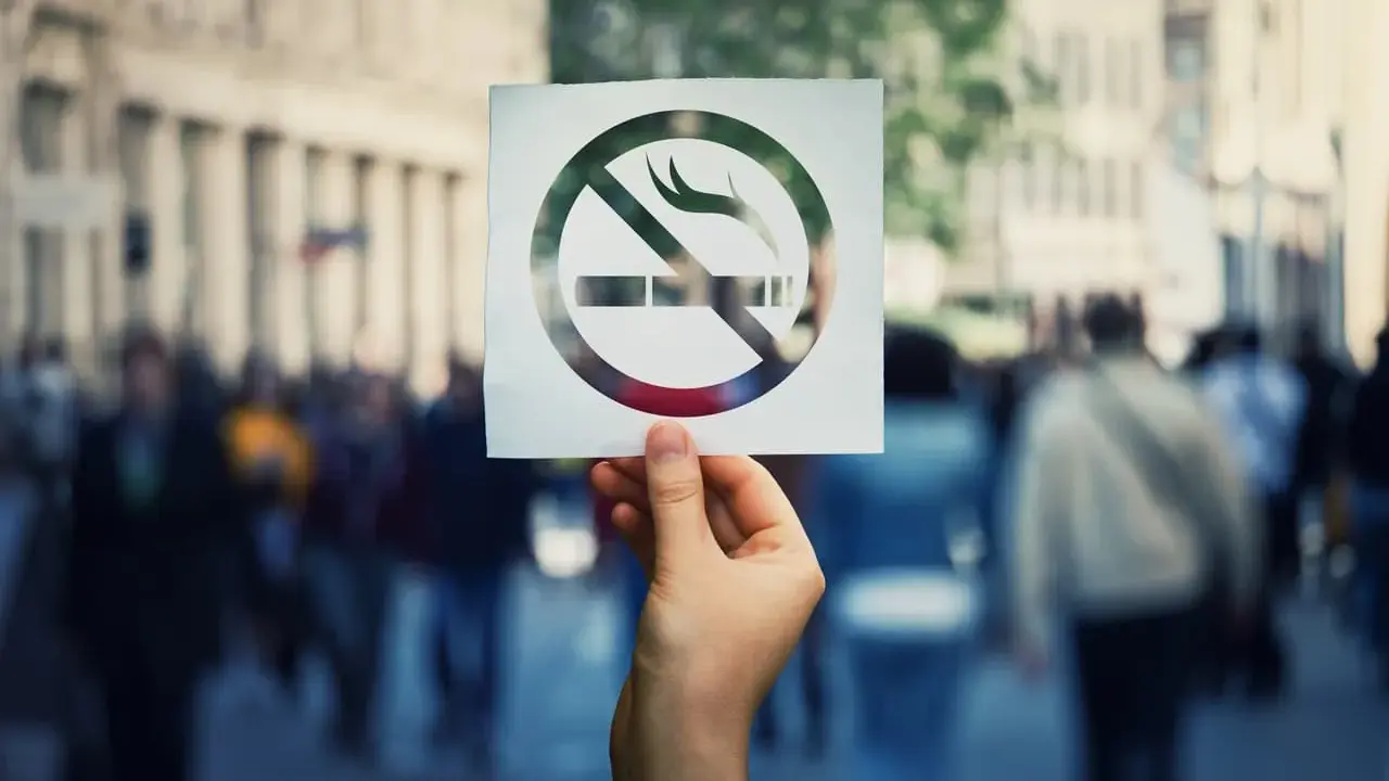 Dłoń trzyma kartkę z symbolem zakazu palenia papierosów na tle tłumu ludzi.