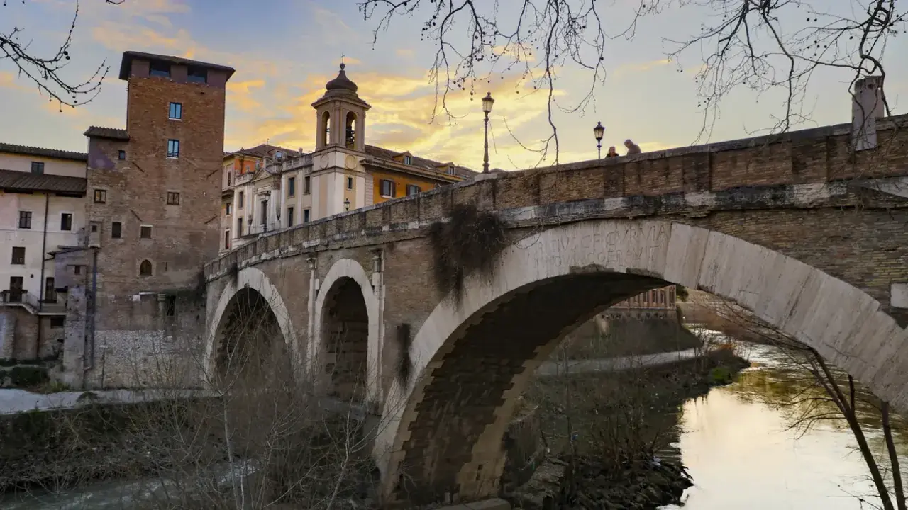 El puente Fabricio, con sus arcos de piedra y la torre medieval, cruza el Tíber bajo un cielo anaranjado.