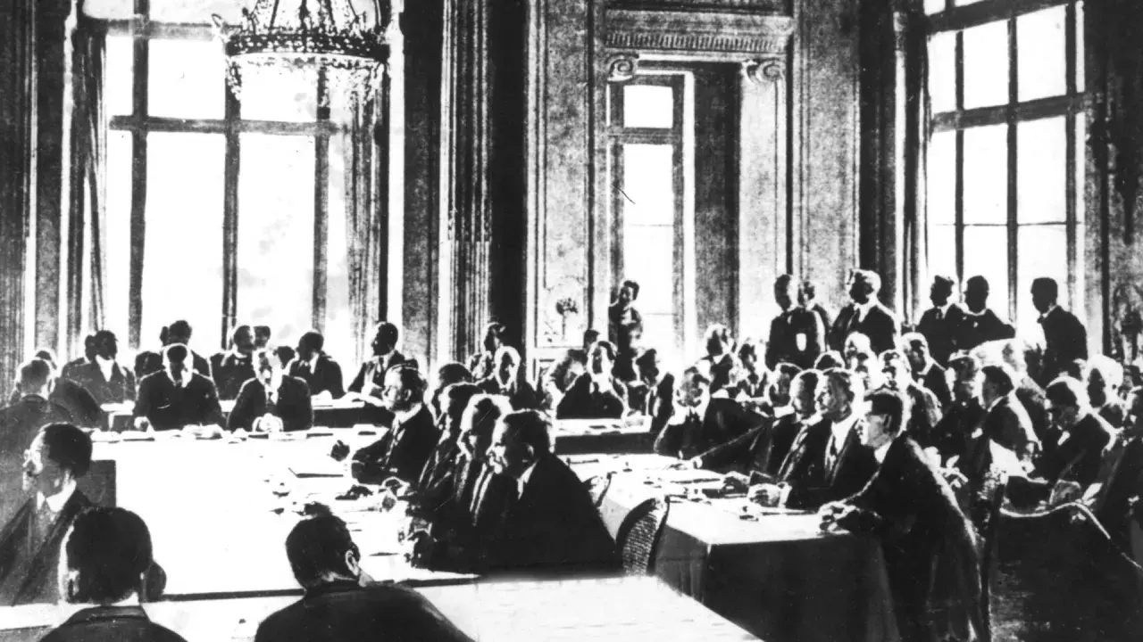 Männer in Anzügen sitzen an einem langen Tisch in einem prunkvollen Saal. Sie diskutieren die Bedingungen des Versailler Vertrags.