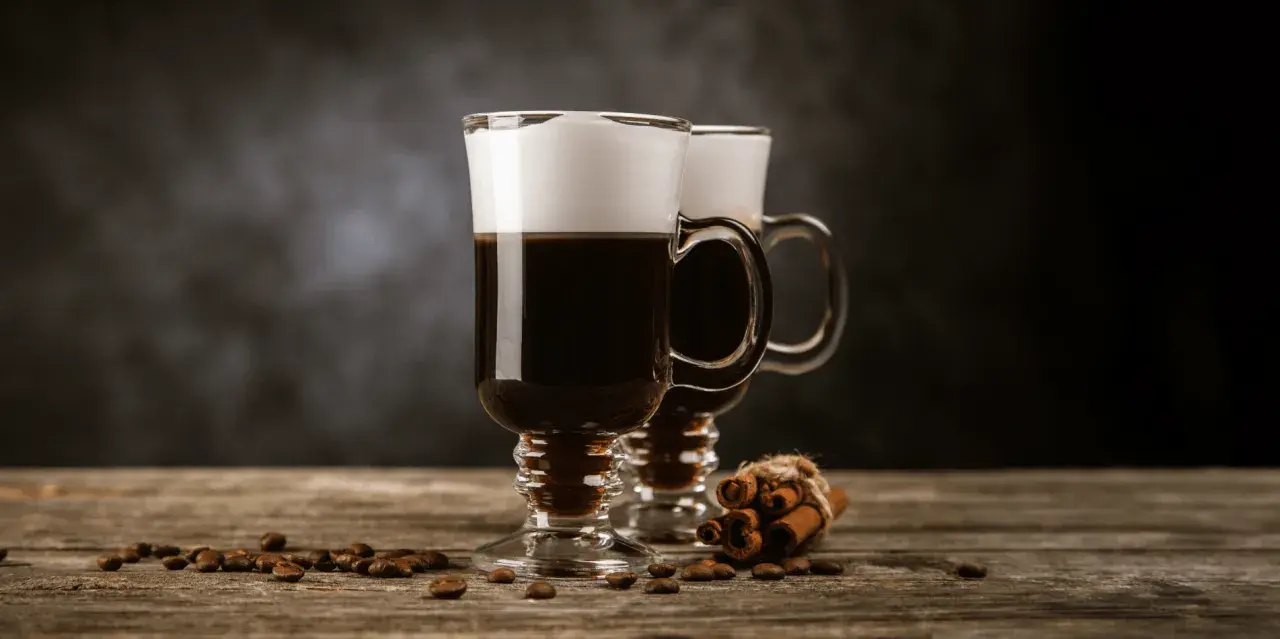 Två glas med kaffe och grädde, kaffebönor och kanelstänger på ett träbord. Perfekt för ostbollar till drinken.