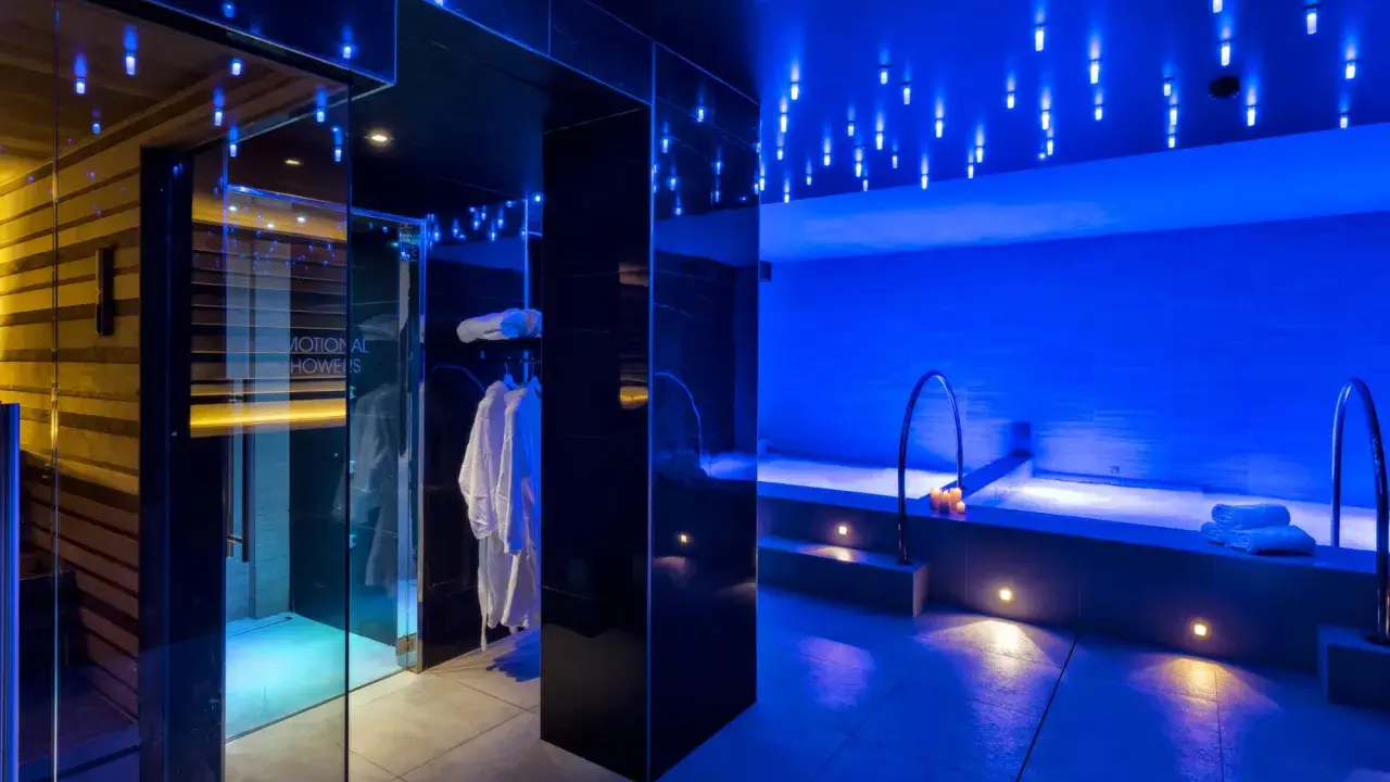 Interni di un centro benessere a Riccione con docce emozionali, vasca idromassaggio e accappatoi bianchi, illuminati da luci blu soffuse.