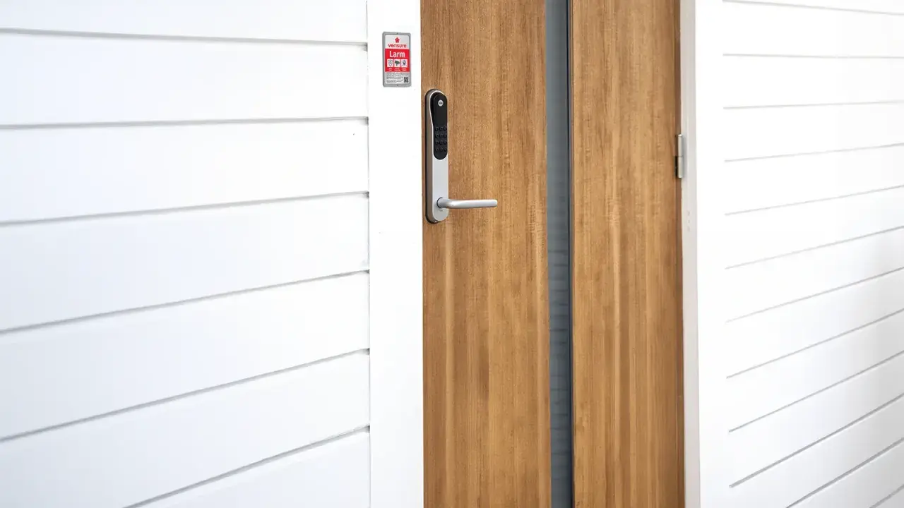 En modern ytterdörr med ett Verisure Yale Doorman V2N lås. Dörren är i trä och har en vitmålad panelvägg bredvid.
