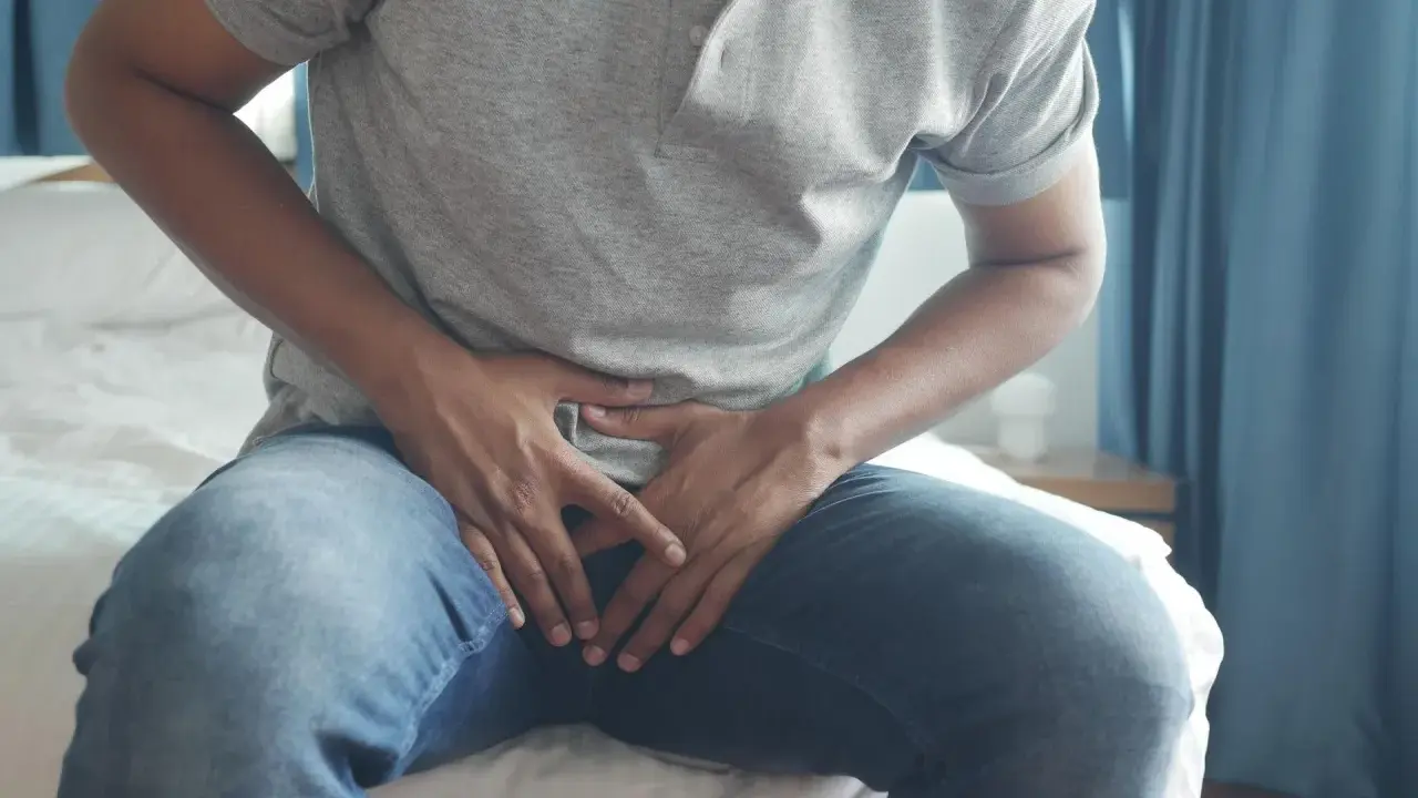 Mężczyzna siedzi na łóżku, trzymając się za podbrzusze. Szuka informacji, jak naturalnie zmniejszyć prostatę.