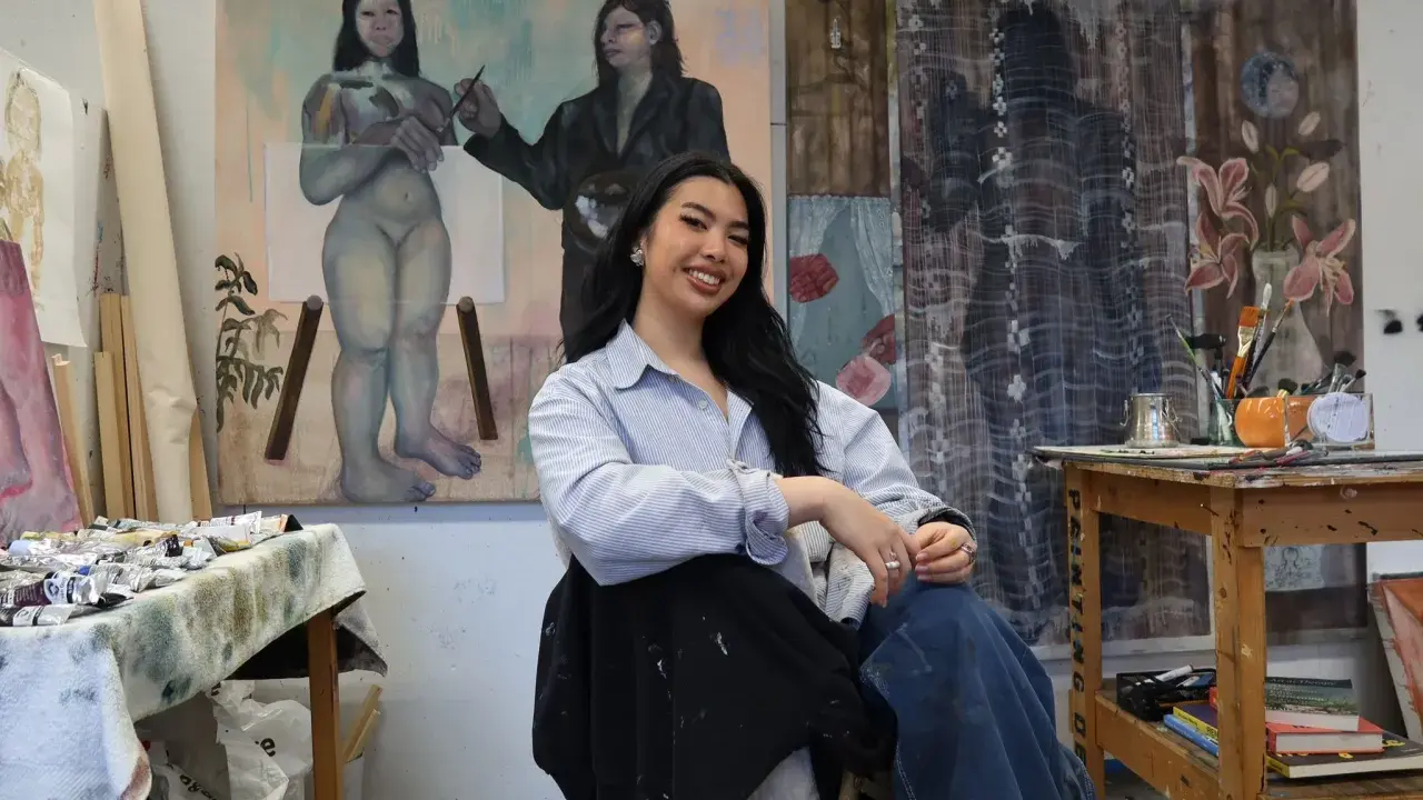 A young woman, one of many talented gen z artists, smiles in her studio, surrounded by her evocative paintings.