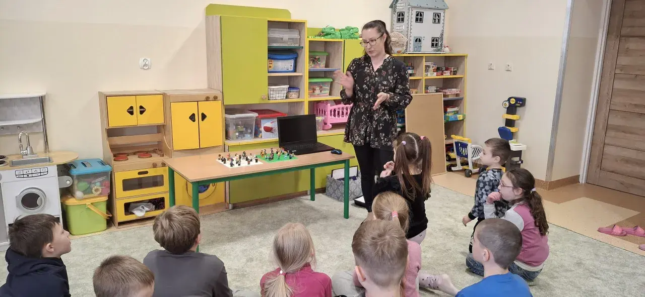 Nauczycielka prowadzi zajęcia kreatywne dla dzieci. Na stole przed nimi leżą klocki Lego, a dzieci z uwagą słuchają.