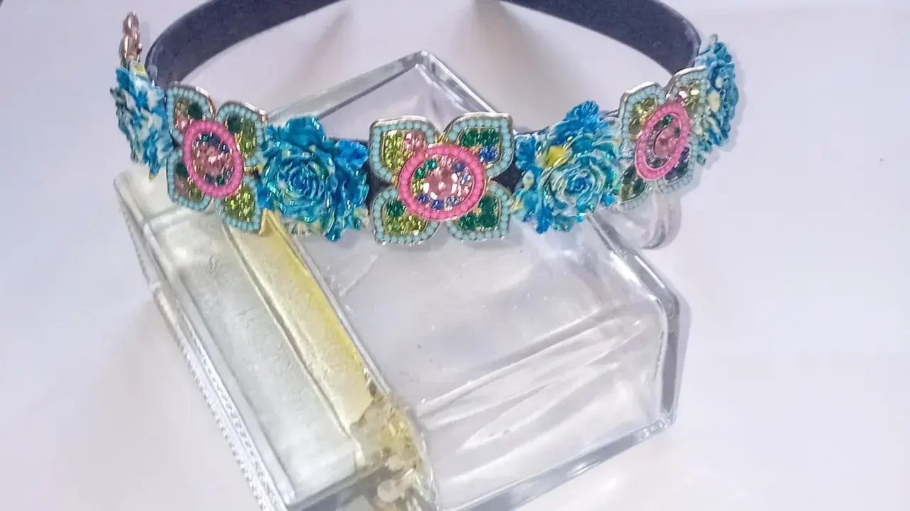 Elegancka opaska z kamieniami w kształcie kwiatów, ozdobiona niebieskimi różami i różowymi detalami, błyszczy na szklanym flakonie.