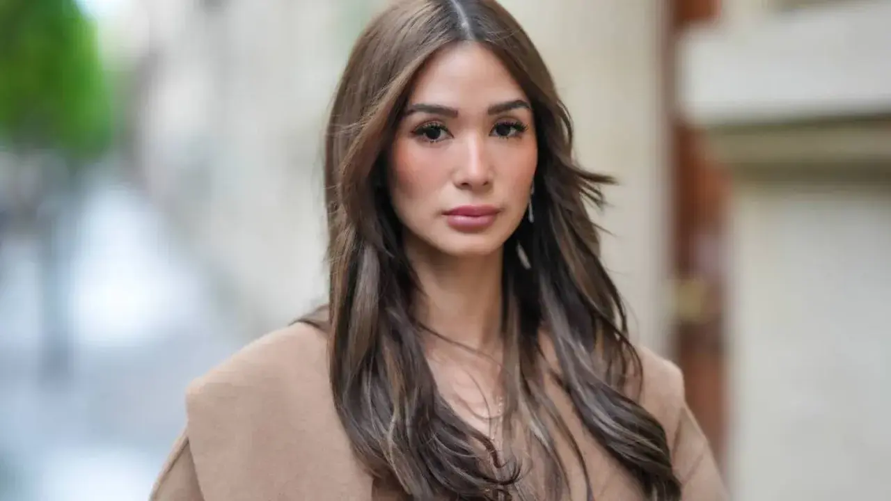 Mujer con cabello castaño largo y ondulado, viste un abrigo beige. Su maquillaje es sutil, un estilo "old money" que realza su belleza natural.