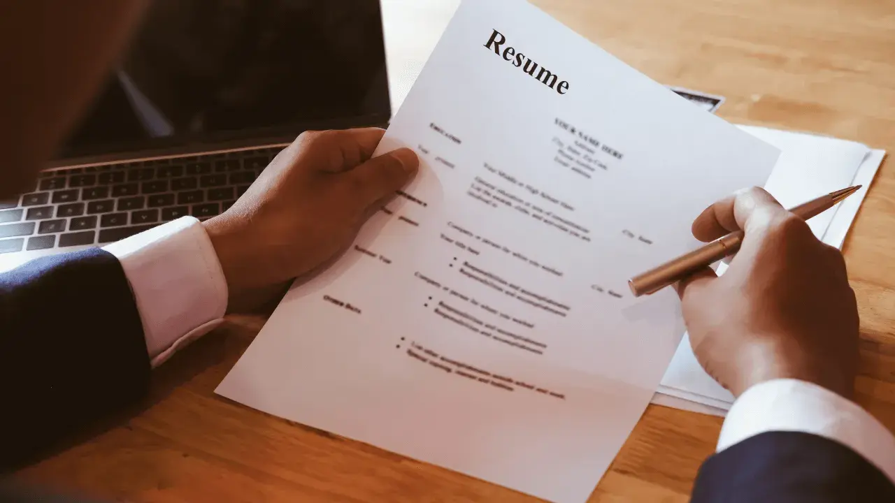 Dłoń trzymająca długopis nad dokumentem CV obok laptopa.