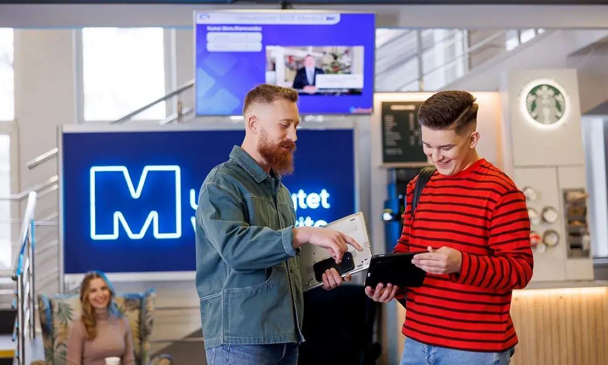 Dwóch studentów dyskutuje o kierunkach studiów WSB, trzymając laptopa i tablet. W tle widoczna jest neonowa litera "M" i ekran informacyjny.