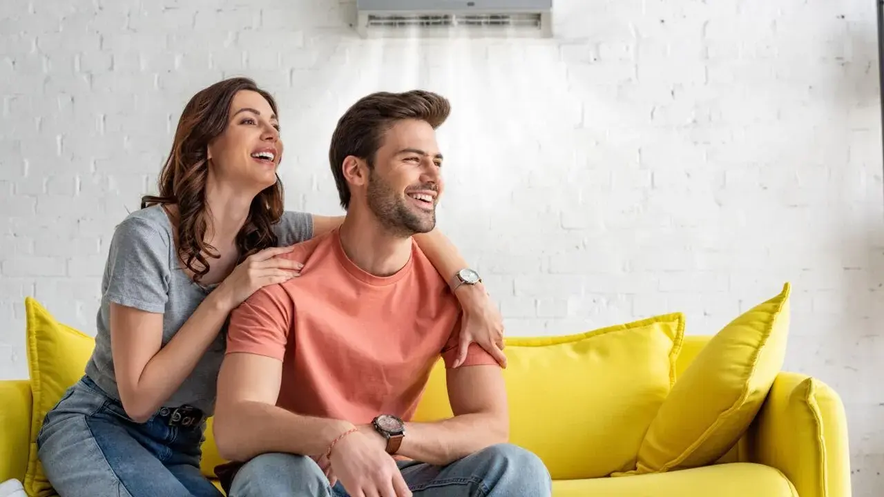 Couple souriant sur un canapé jaune, profitant de la brise fraîche d'un climatiseur moins de 50 db.