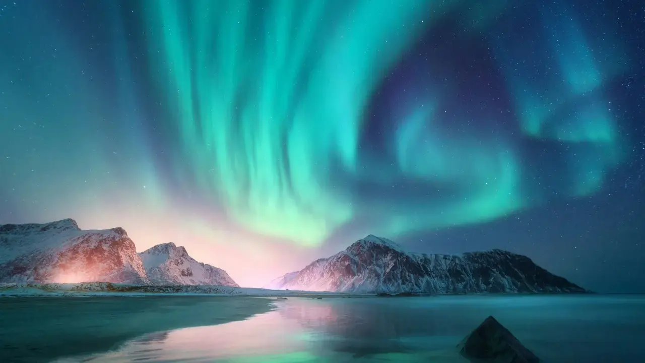 En magisk aurora himmel över snöklädda berg och en spegelblank strand. Norrskenet dansar i gröna och lila nyanser.