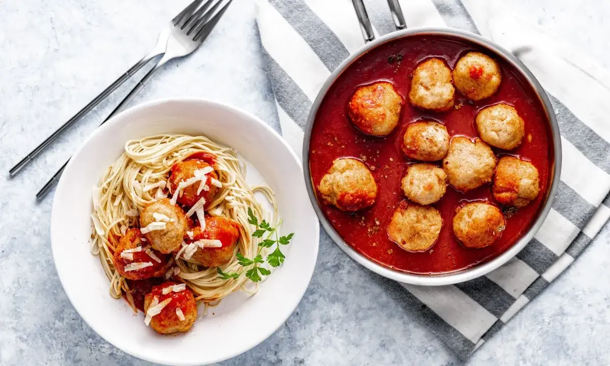 Pyszne pulpety w sosie pomidorowym z makaronem spaghetti, posypane parmezanem. Obok patelnia z gorącymi pulpetami w sosie.