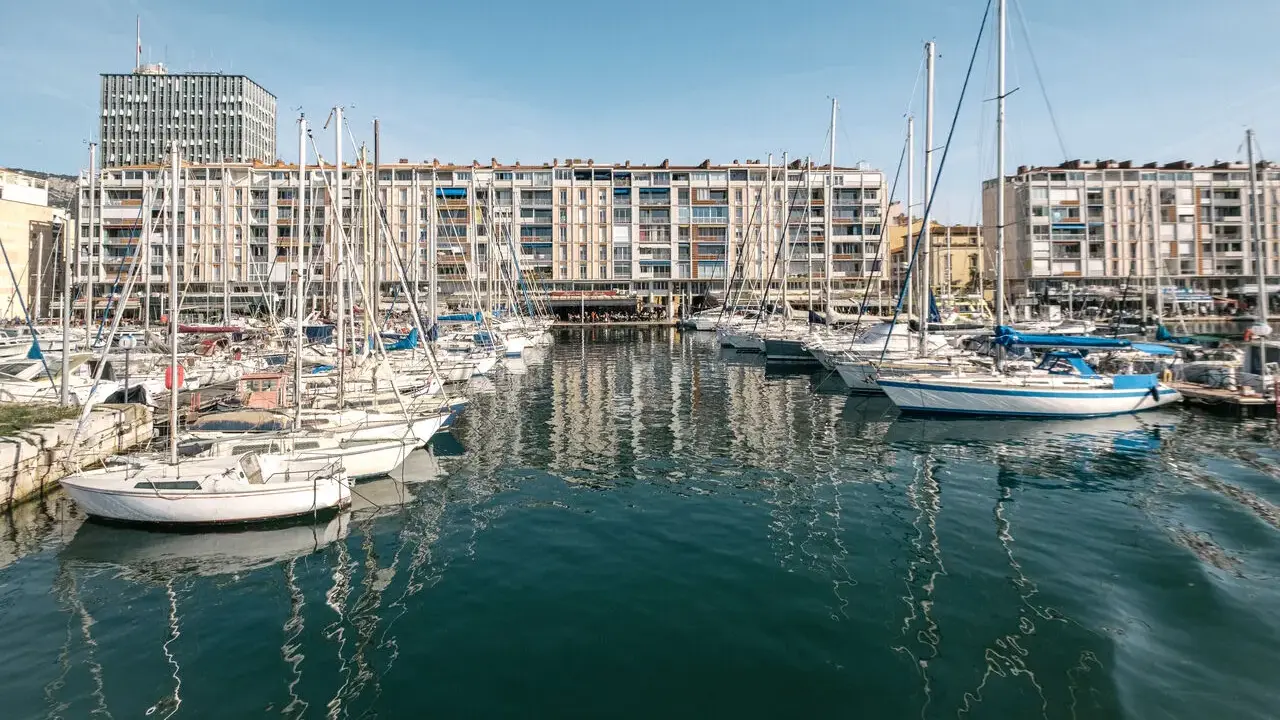 Barche a vela ormeggiate nel porto di Tolone, con edifici residenziali sullo sfondo e il cielo azzurro.
