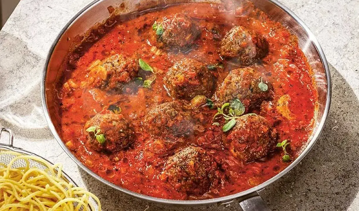 Frikadeller i tomatsås som puttrar i en panna, med en sidokorg med spaghetti.