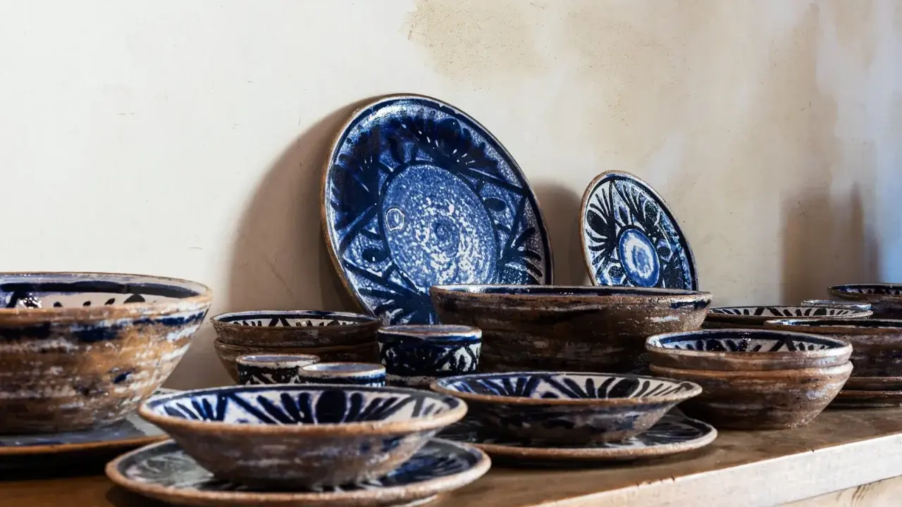 Ceramiche artigianato toscano con decori blu su fondo grezzo. Piatti e ciotole disposti su una mensola di legno.