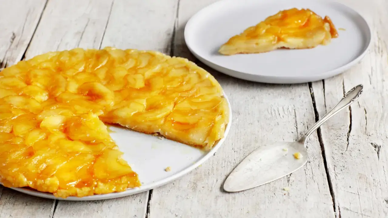Eine goldbraune Tarte Tatin mit karamellisierten Äpfeln auf einem Teller, daneben ein Stück auf einem weiteren Teller und ein Tortenheber.