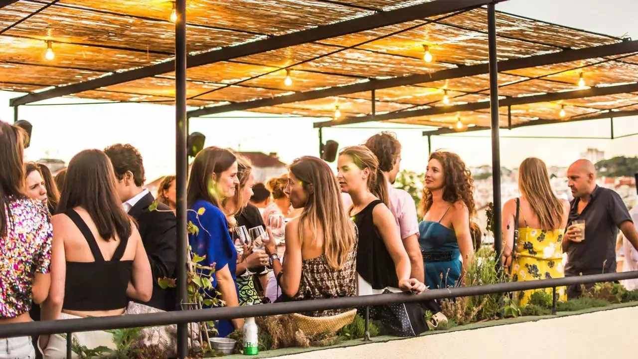 Menschen genießen den Abend auf einer Dachterrasse in einem **bar lissabon**. Unter einem Strohdach mit Lichterketten unterhalten sie sich bei Getränken.