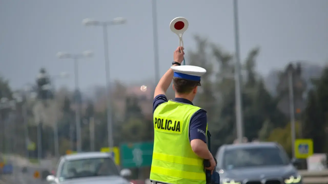Policjant kieruje ruchem, podnosząc znak stop. W tle widać samochody i słupy oświetleniowe, sugerujące podwyższone **stopnie alarmowe**.