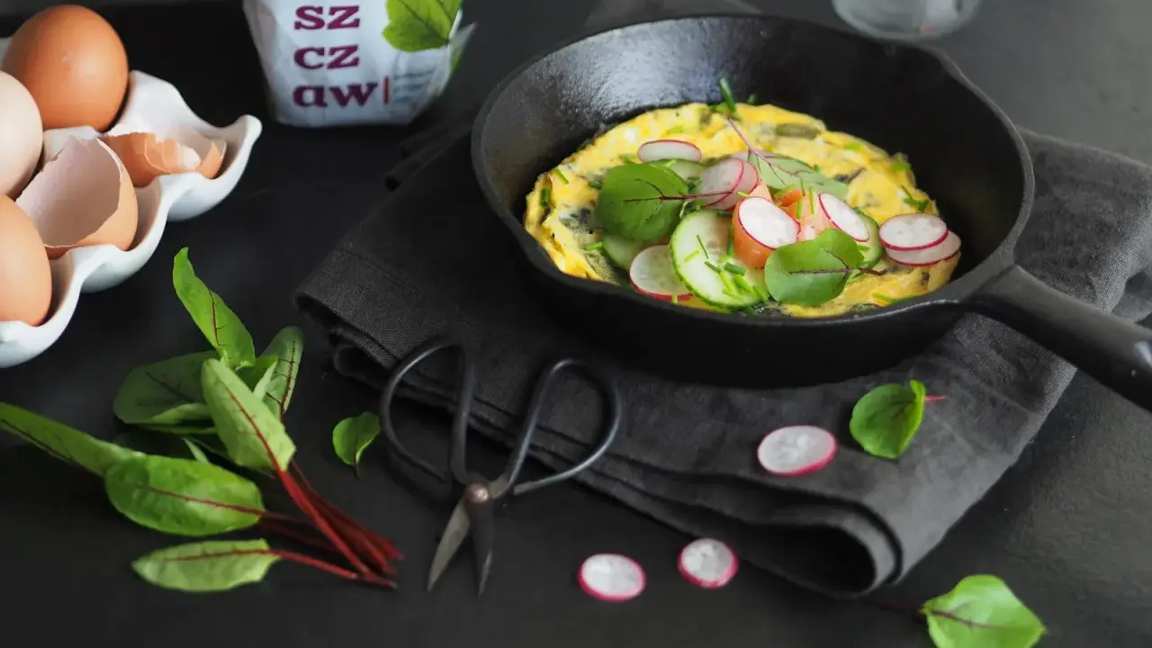 Jak zrobić dobry omlet? Oto inspiracja: puszysty omlet z rzodkiewką, ogórkiem i ziołami na czarnej desce.