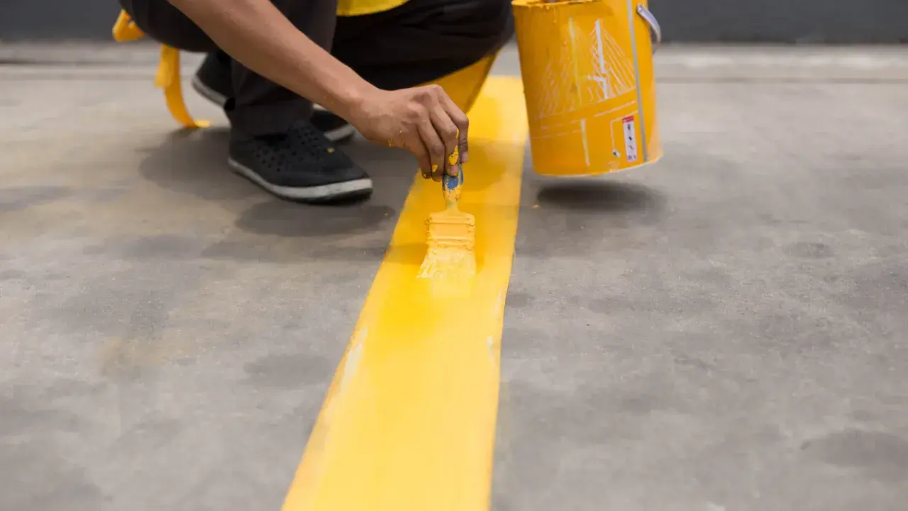 Persona pintando una línea amarilla sobre suelo de cemento sin pulir, usando una brocha y un cubo de pintura.