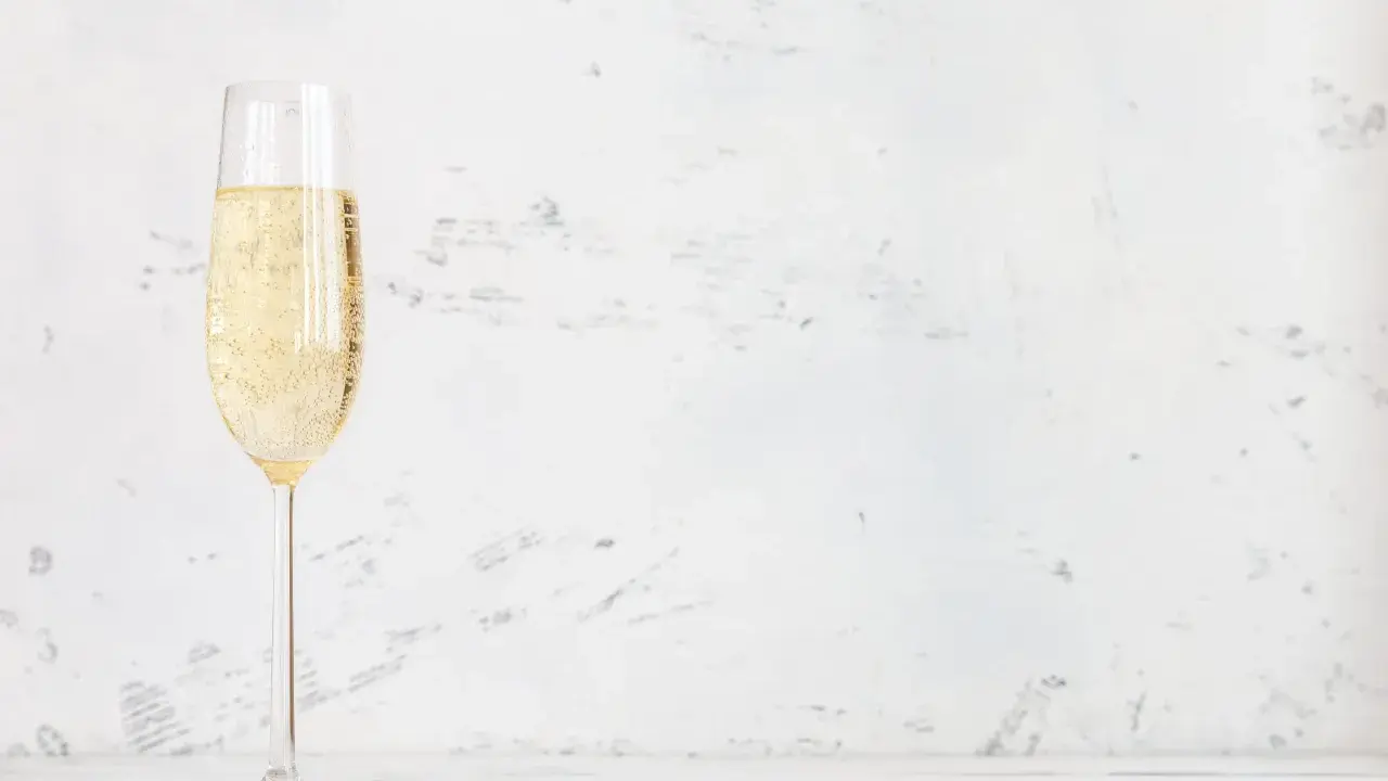 Ett glas champagne med bubblor, kanske en Jaques Selosse, står på ett vitt bord mot en ljus, texturerad bakgrund.