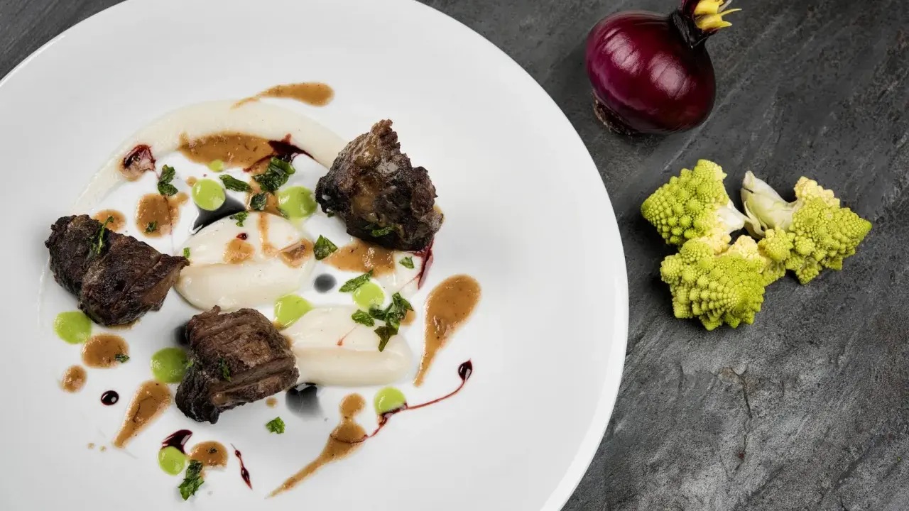 Succulenta guancia di manzo tenerissima, servita su purè bianco con gocce verdi e salsa bruna. Accanto, cipolla rossa e cavolfiore romanesco.