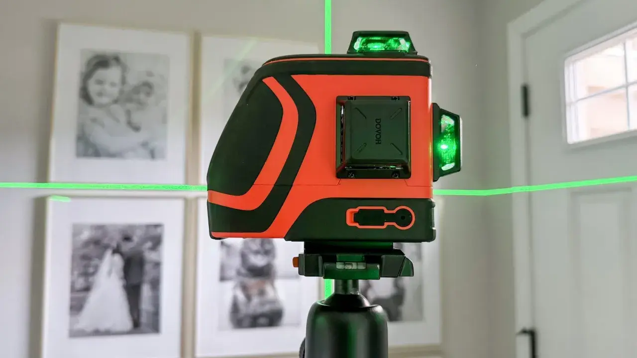 Pomarańczowa poziomica laserowa z zielonymi liniami na statywie, na tle oprawionych zdjęć.