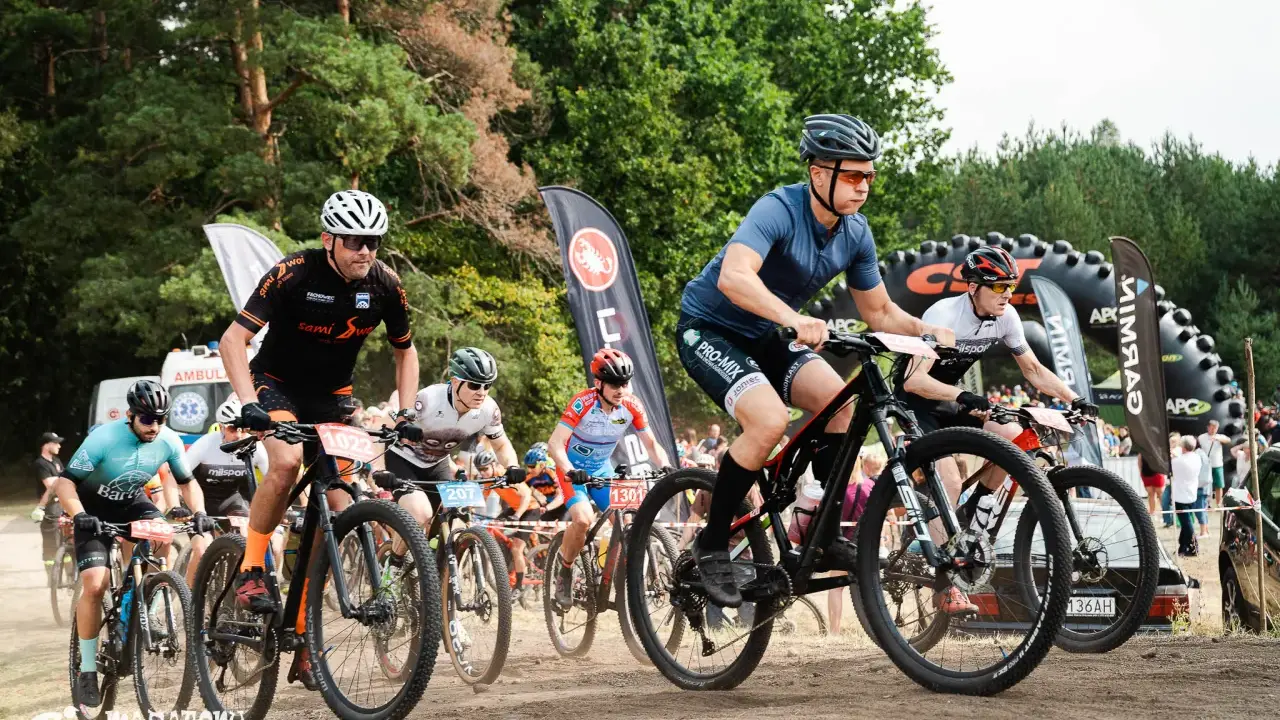 Kolarze biorą udział w wyścigu **zawody MTB**. Widać ich w akcji, jadących po piaszczystej trasie, z lasem w tle.